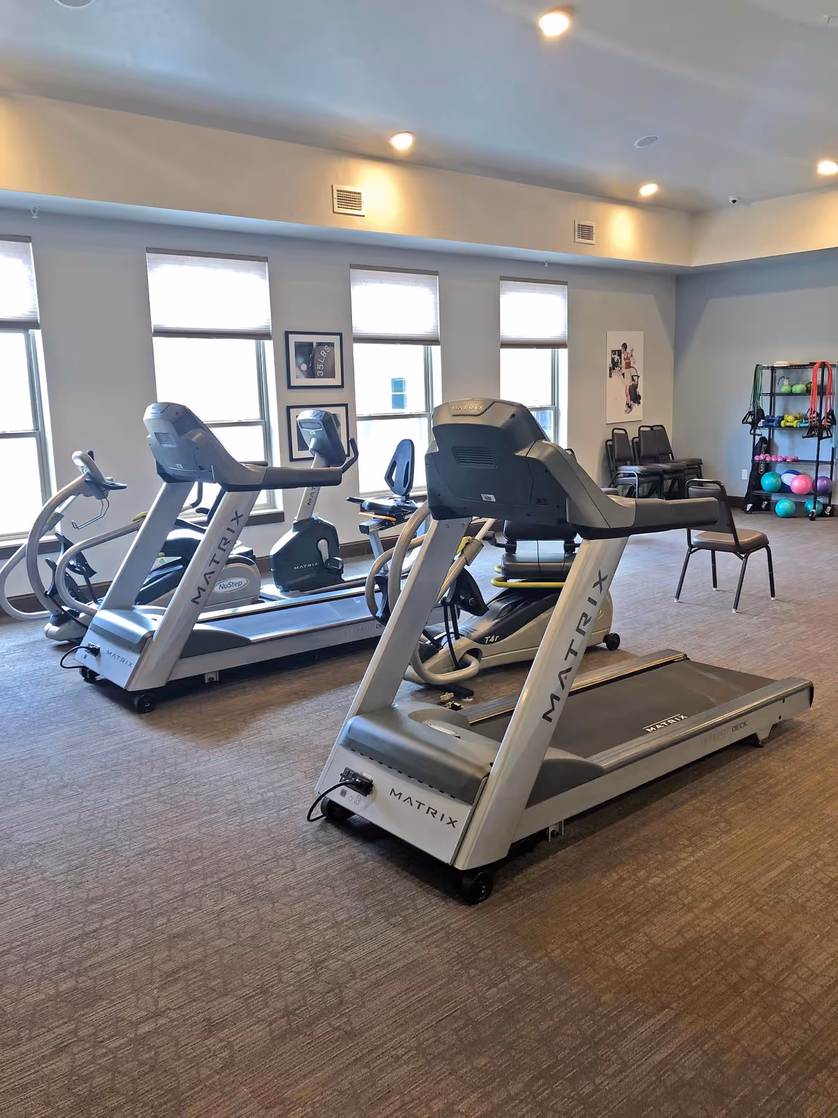 A fitness room with two treadmills, an exercise bike, and various exercise balls and resistance bands on a rack. The room has large windows with blinds, carpeted floor, and recessed ceiling lights.