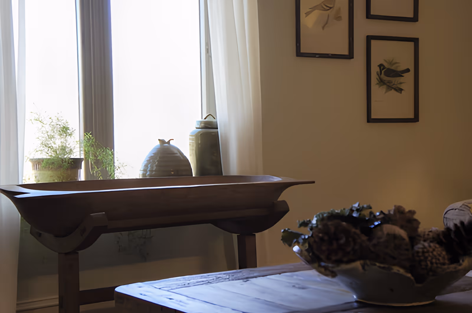 A cozy interior corner with a wooden table in the foreground holding a decorative bowl filled with pine cones and greenery. Behind it, a wooden bench or tray table is positioned in front of a window with sheer white curtains. On the windowsill, there are two ceramic vases and a potted plant. The wall to the right features framed bird illustrations.