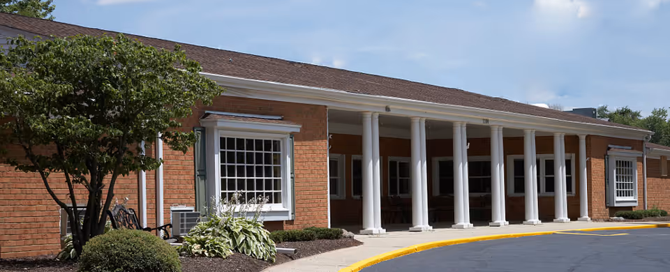 Brick single-story building entrance with white columns, large windows, landscaping, and a curved driveway.