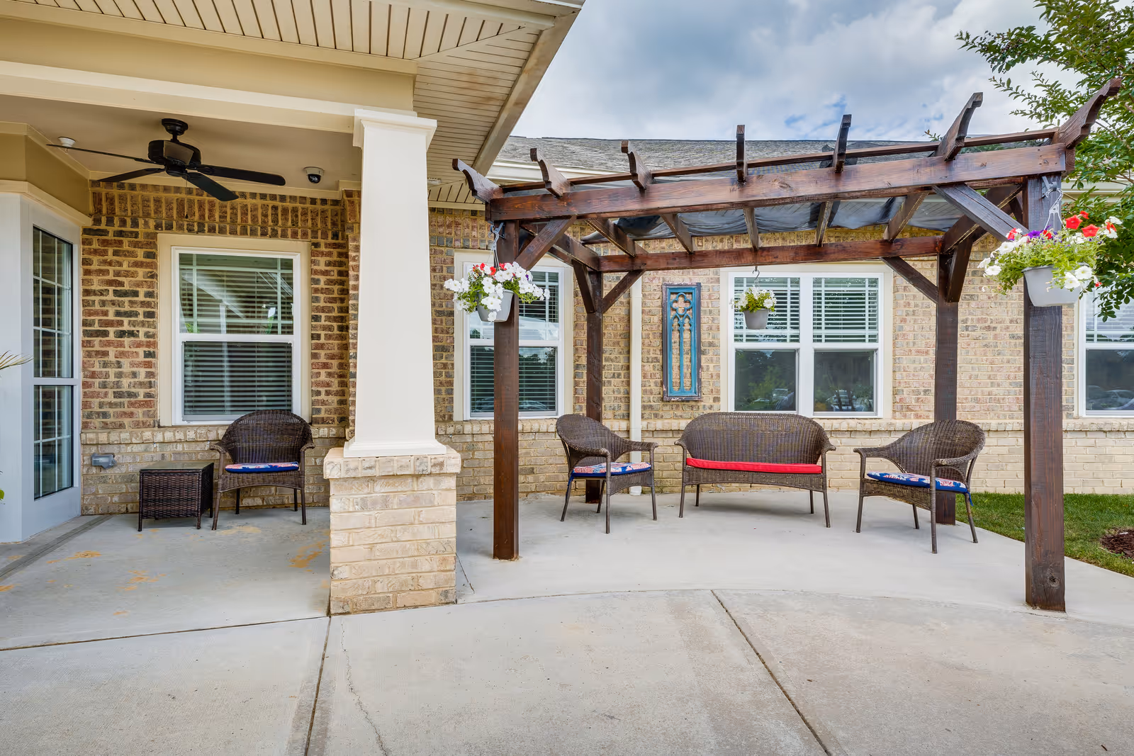 Outdoor seating area at Chatham Ridge Assisted Living featuring a wooden pergola with hanging flower pots, wicker chairs with cushions, and a brick building facade with windows in the background.