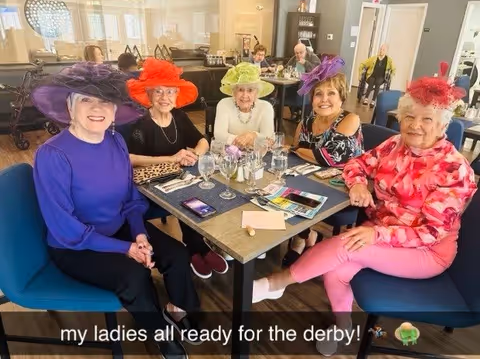 Five elderly women sitting around a dining table in a senior living facility, each wearing colorful, decorative hats. The table is set with glasses, napkins, and some papers. Other residents and staff are visible in the background in a bright, spacious room.