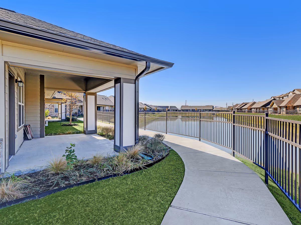 Covered patio area with concrete flooring and columns, adjacent to a curved concrete walkway bordered by a black metal fence overlooking a pond. Residential buildings are visible in the background under a clear blue sky.