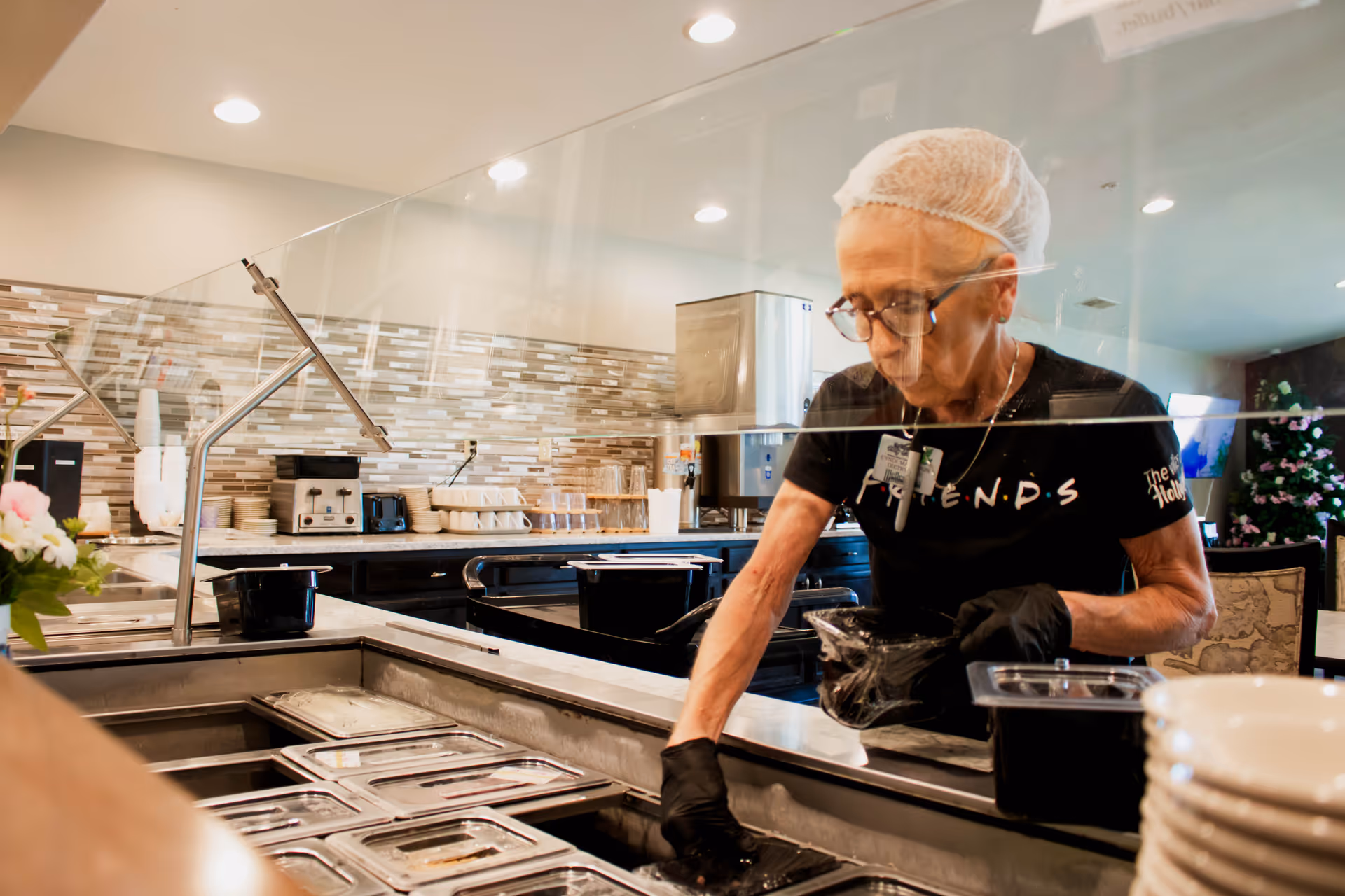 A staff member wearing a hairnet and gloves serves meals behind a cafeteria-style counter with food trays and a glass sneeze guard.
