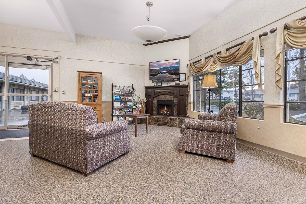 A cozy senior living facility seating area with two patterned armchairs facing a fireplace and wall-mounted TV, large windows, and a small table and bookshelf.