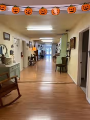 A long hallway in a senior living facility decorated with a string of pumpkin-shaped Halloween decorations hanging from the ceiling. The hallway has wooden flooring and is lined with chairs, small tables, lamps, and framed pictures on the walls. Doors to rooms are visible on both sides, and natural light is coming from windows at the far end.