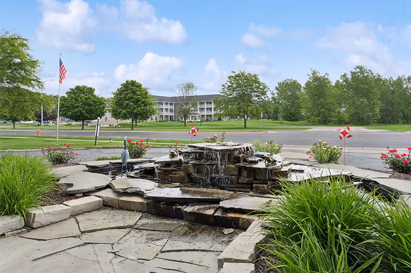 Outdoor view of a senior living facility with a stone water fountain in the foreground, surrounded by green plants and flowers. In the background, there is a large building with multiple balconies, trees, and an American flag on a flagpole under a partly cloudy sky.