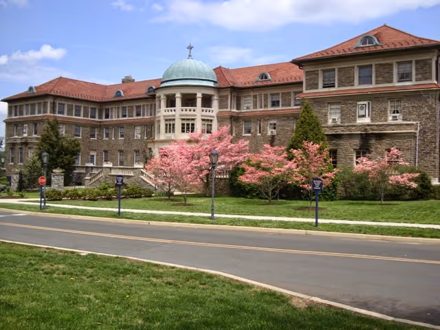 Stone three-story building with a central domed cupola topped by a cross, red tile roof, pink flowering trees and a lawn beside a road.