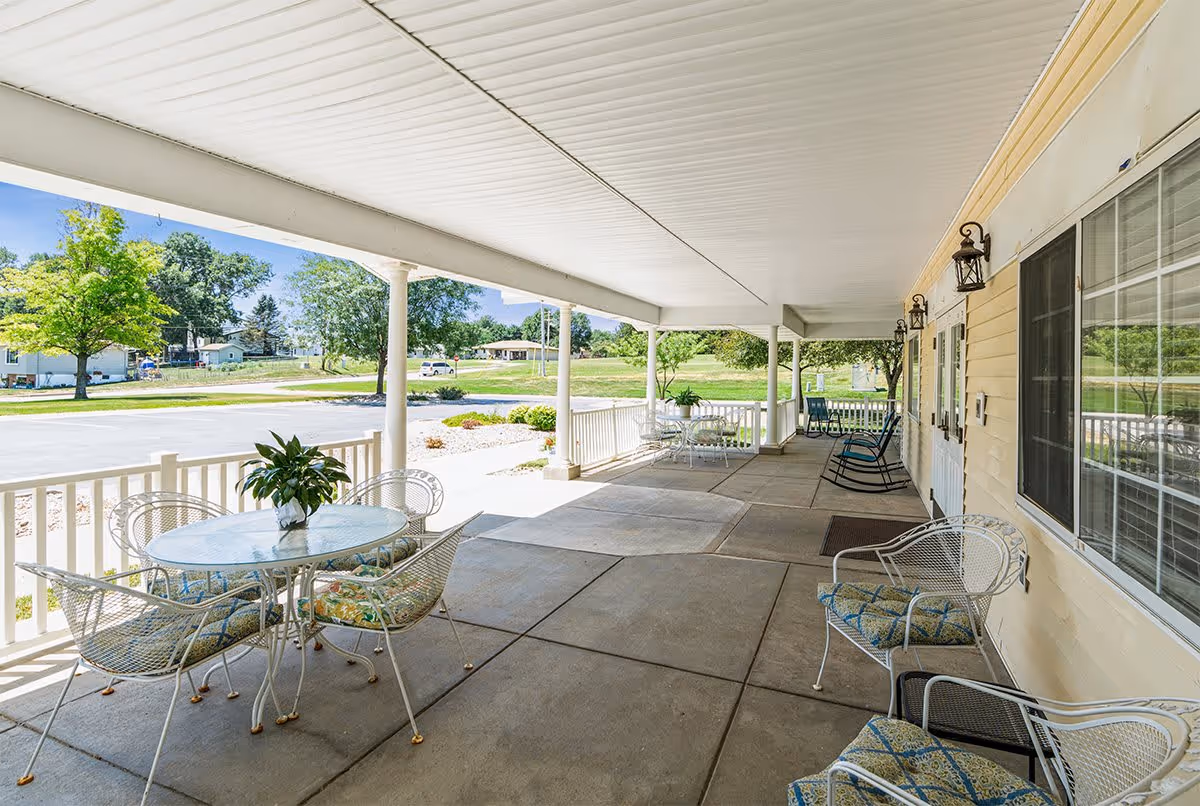 Covered outdoor patio area with white metal tables and chairs with cushions, potted plants on tables, rocking chairs along the wall, and a view of a parking lot and green trees in the background.