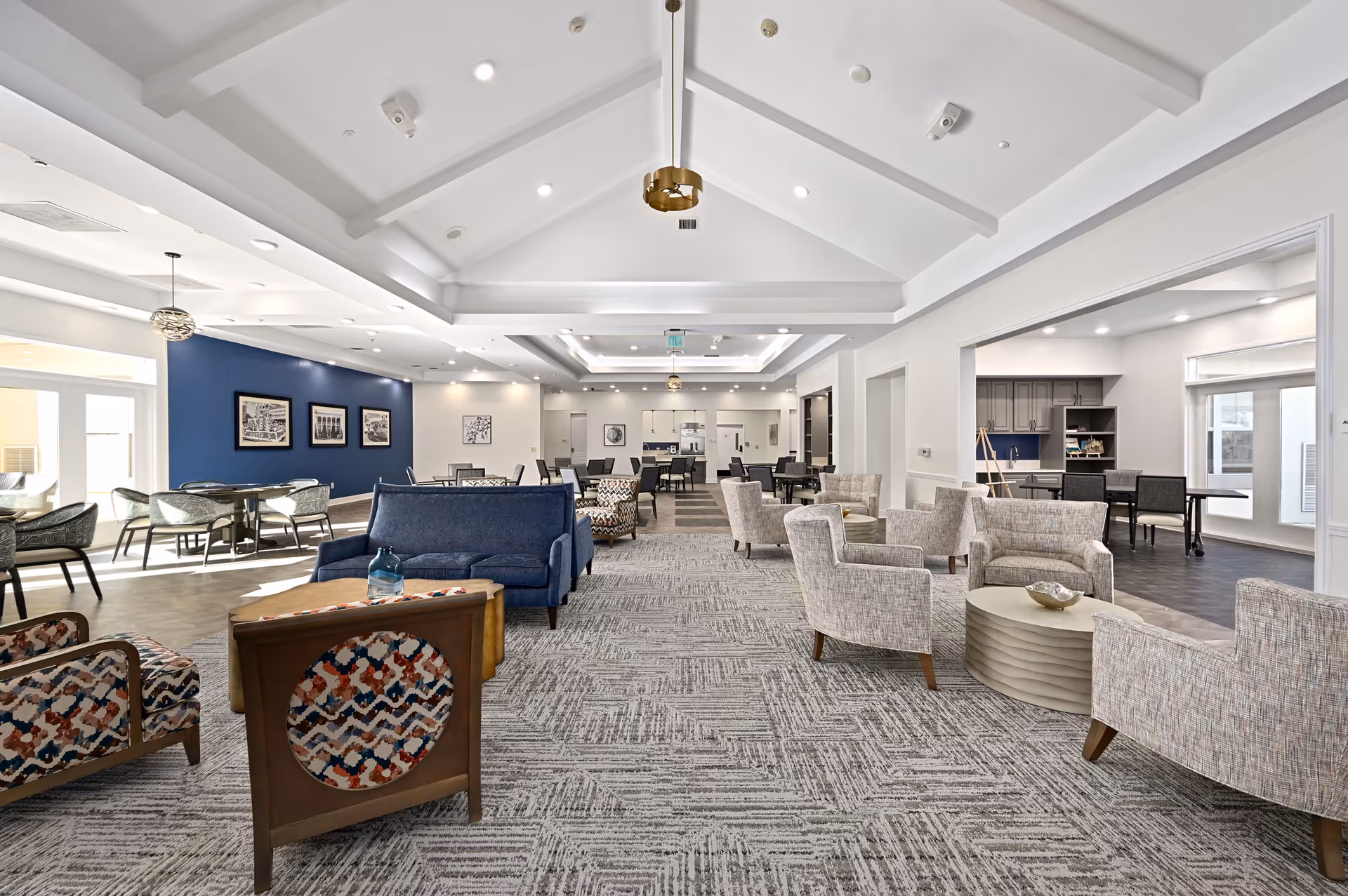 A spacious and well-lit senior living common area with a high vaulted ceiling and modern lighting fixtures. The room features a mix of comfortable seating including patterned armchairs, a blue sofa, and beige chairs arranged around circular tables. In the background, there are dining tables and chairs near a blue accent wall with framed artwork. To the right, there is an open area with a kitchenette and additional seating. The flooring combines carpet and wood-like materials.
