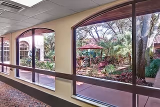View through large arched windows from an indoor hallway looking out onto a garden area with trees, plants, a red umbrella, and outdoor seating.