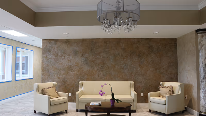 A cozy sitting area with a beige sofa and two matching armchairs arranged around a dark wooden coffee table with a purple orchid plant and a magazine on it. The background features a textured brown wall and a chandelier hanging from the ceiling. Large windows on the left side allow natural light into the space.