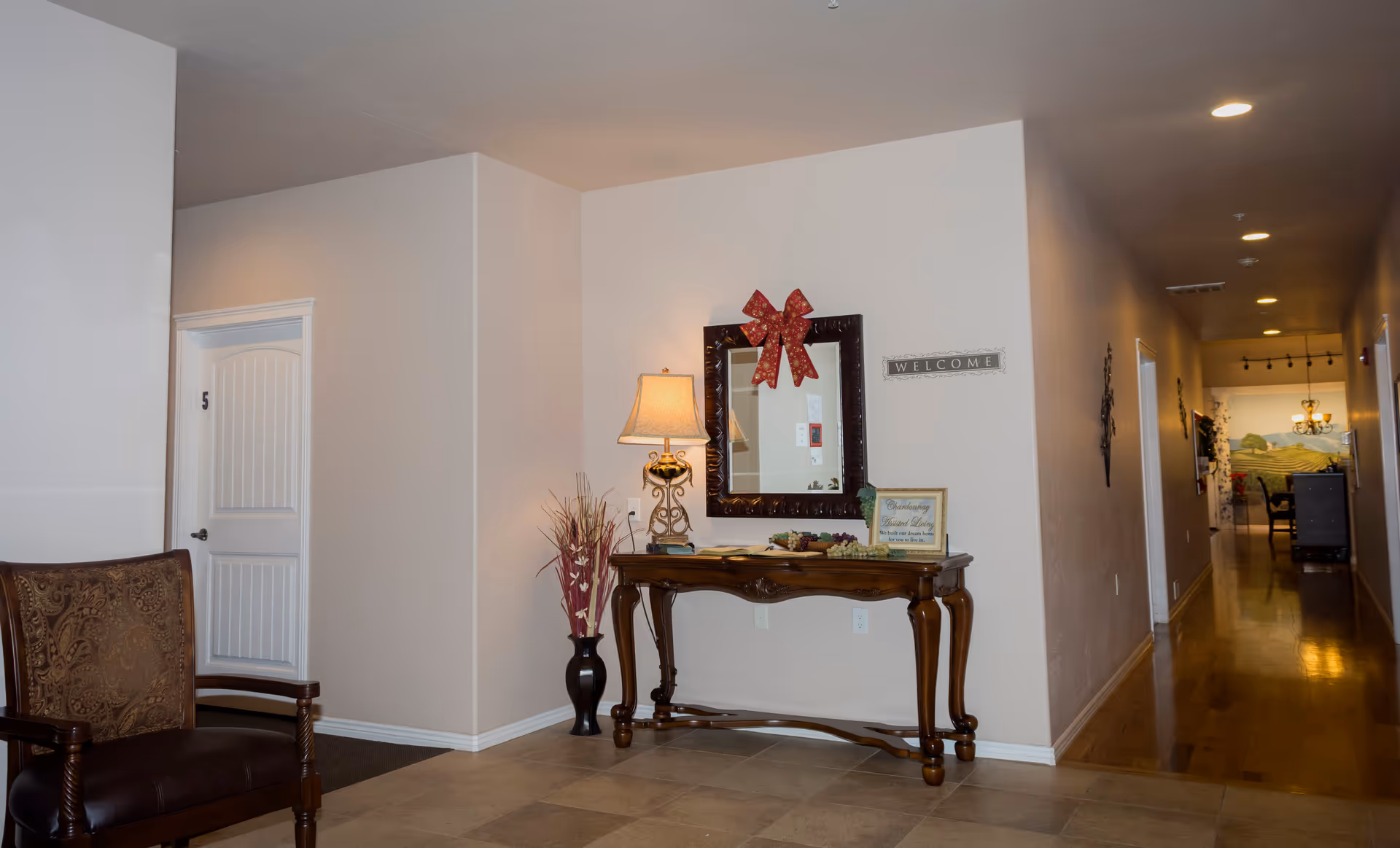 Interior hallway of an assisted living facility with a wooden chair on the left, a wooden console table with a lamp, decorative vase, and a mirror with a red bow on the wall above it. A 'WELCOME' sign is mounted on the wall next to the mirror. The hallway extends to the right with doors and a mural visible in the distance.