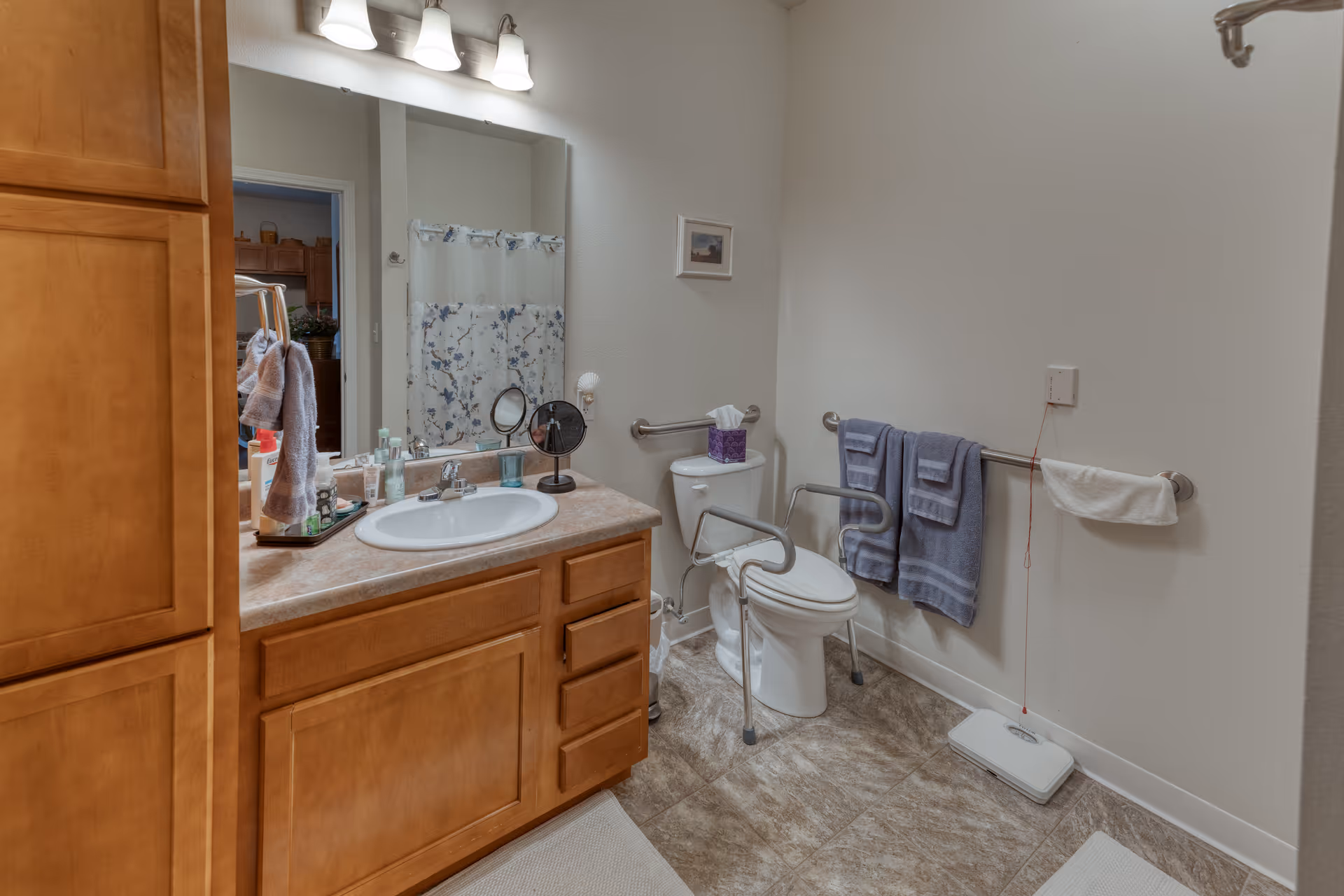 A bathroom with a wooden vanity and sink on the left, a large mirror above the sink with three light fixtures, a toilet with safety rails, and two blue towels hanging on a grab bar on the right wall. There is a white scale on the floor and a shower curtain with a floral pattern visible in the mirror reflection.