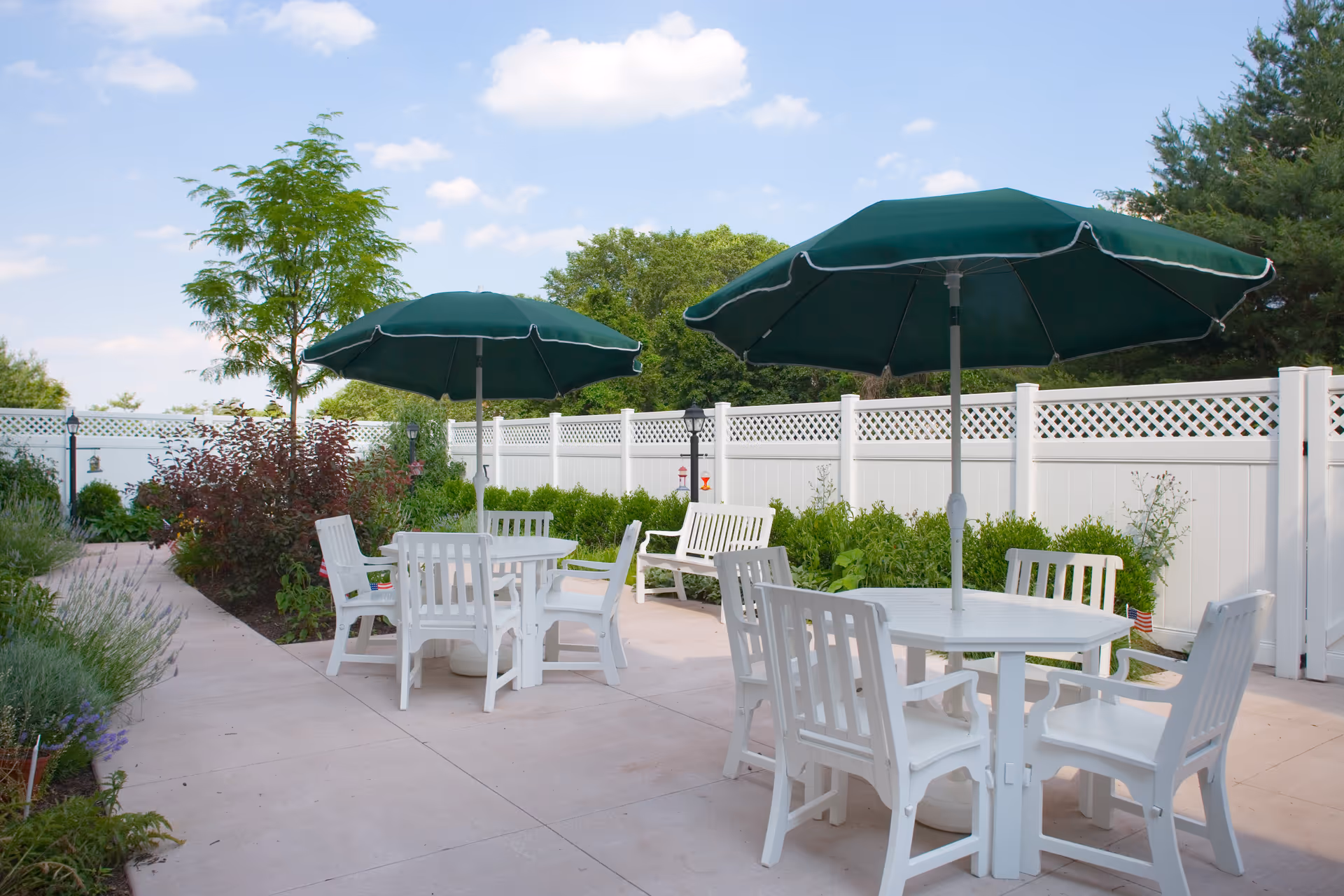 Outdoor patio with white tables and chairs under green umbrellas beside a white lattice fence.