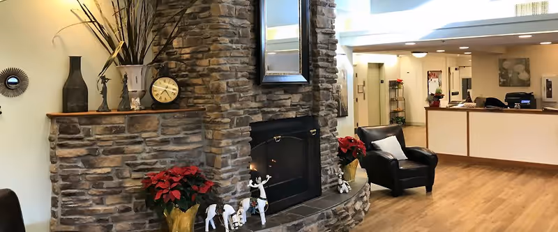 A cozy assisted living facility interior featuring a stone fireplace decorated with a clock, vases, and holiday figurines. A black leather armchair with a white pillow is positioned near the fireplace. In the background, there is a reception desk with a computer and some decorations, and a hallway leading to other rooms.
