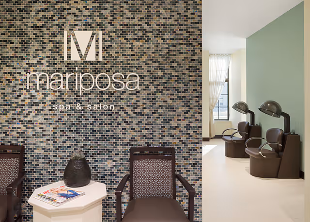 Interior view of Mariposa spa & salon with two patterned chairs and a small white table holding a decorative vase and magazines in front of a mosaic tiled wall. In the background, there are three brown salon hair dryers with chairs underneath near a window with sheer curtains.