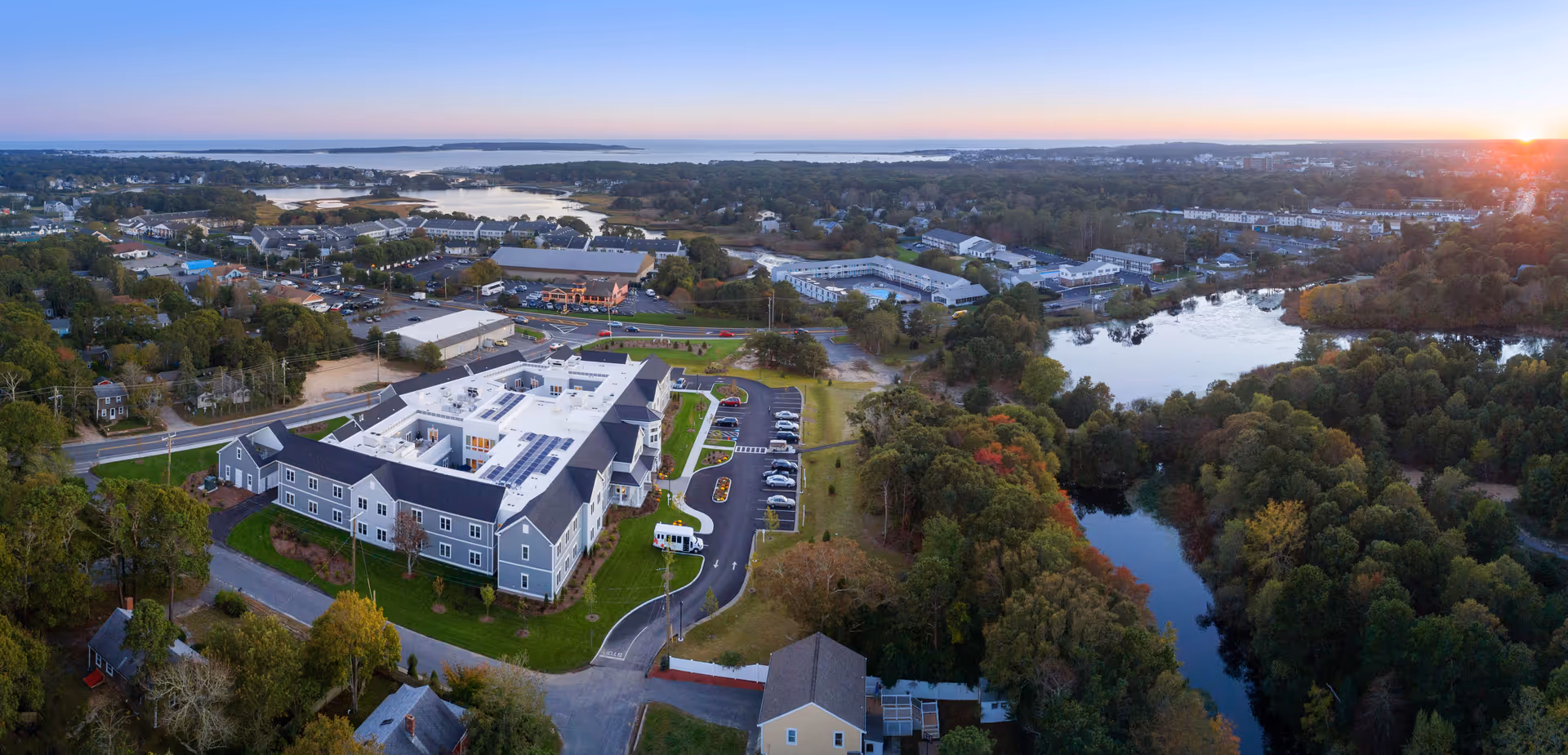 Aerial view of a senior living facility named Maplewood at Mill Hill surrounded by trees, water bodies, and a small town. The building is large with a parking lot and roads nearby, set against a sunset sky.