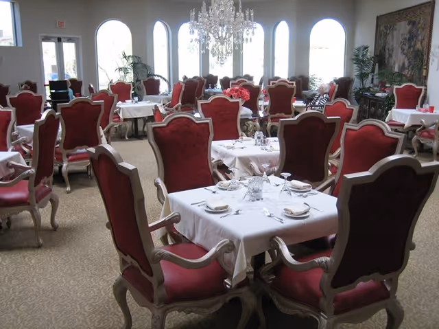 Elegant dining room with multiple tables covered in white tablecloths, set with plates, napkins, glasses, and silverware. The chairs are upholstered in red fabric with ornate wooden frames. Large arched windows let in natural light, and a crystal chandelier hangs from the ceiling. There are plants and a decorative tapestry on the walls.