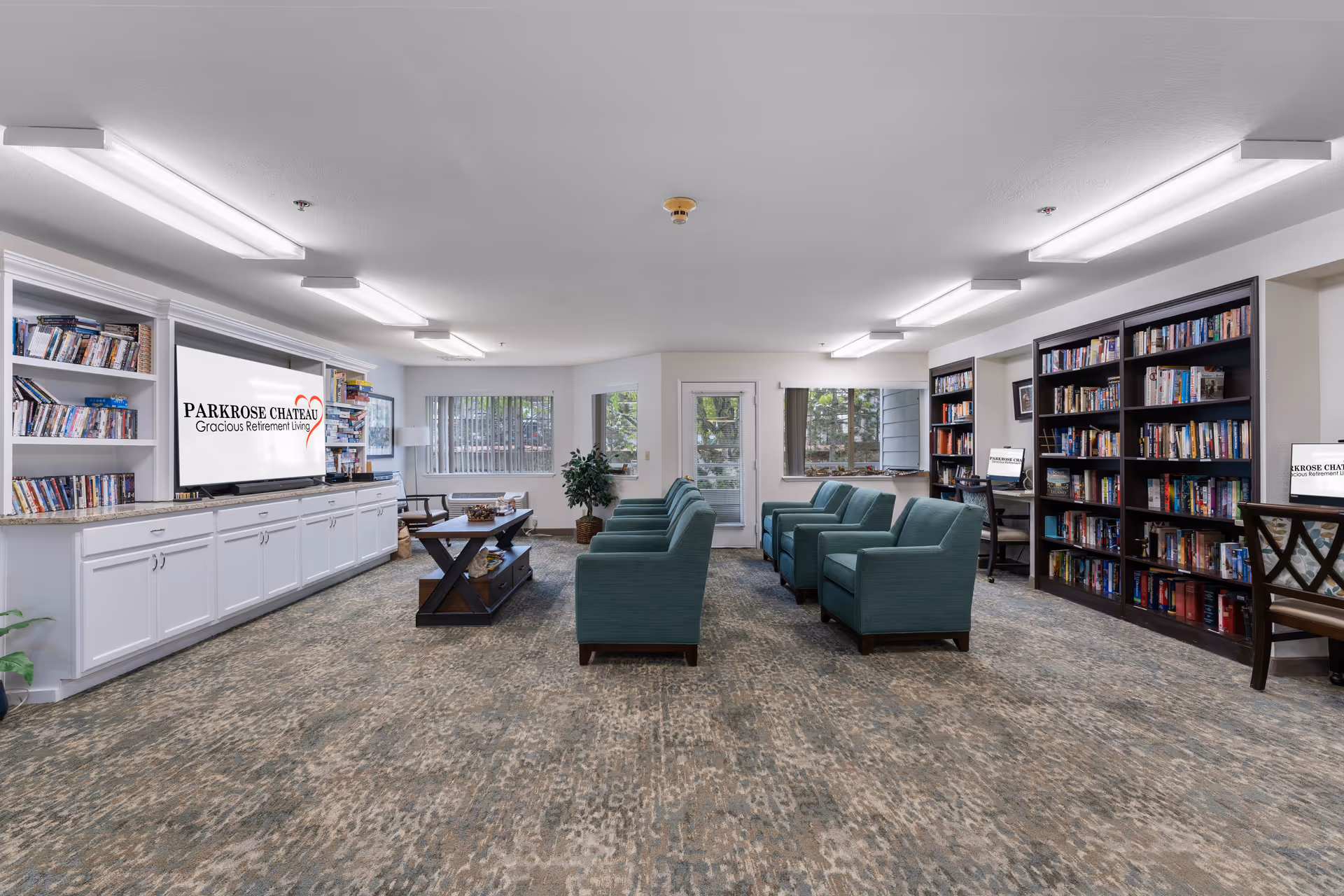 A spacious and well-lit common area in a retirement living facility featuring multiple teal armchairs arranged in rows facing a large TV screen displaying the Parkrose Chateau Gracious Retirement Living logo. The room has built-in white cabinets and shelves filled with books and DVDs, a coffee table in the center, and large windows allowing natural light to enter.