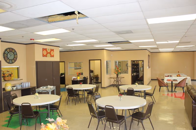 Community dining/activity room with several round tables and chairs, a kitchenette and office doorways beneath a dropped ceiling with a missing tile.