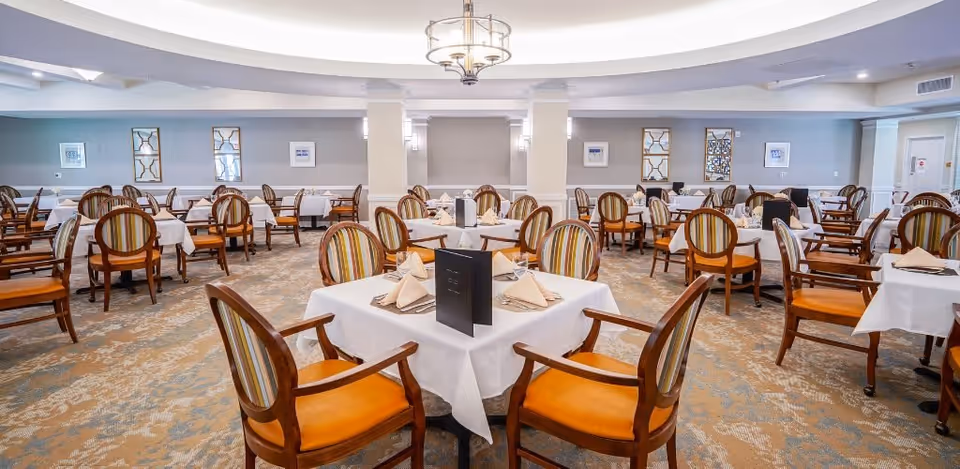 A spacious dining room with multiple tables covered in white tablecloths, each set with folded napkins and menus. The chairs have wooden frames with orange seats and striped backs. The room has soft lighting, decorative wall art, and a patterned carpet.
