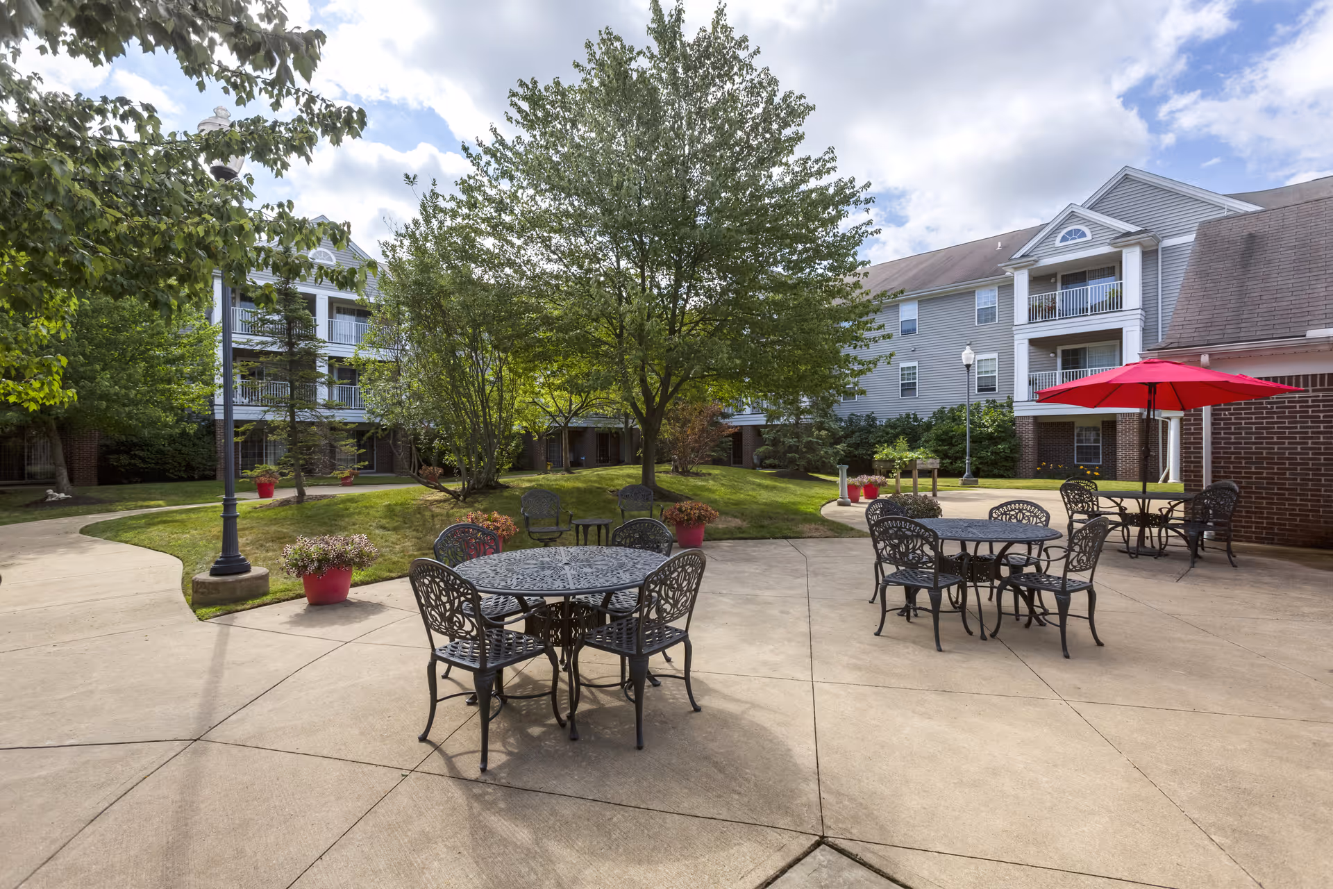 Outdoor patio area at Independence Village of Avon Lake with metal tables and chairs, some with red umbrellas, surrounded by trees and greenery, with a multi-story residential building in the background under a partly cloudy sky.
