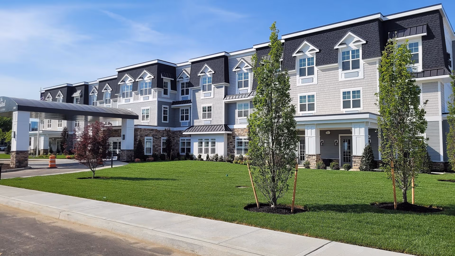 Front exterior of a three-story assisted living building with a covered entrance, manicured lawn, and young trees.