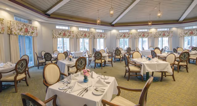 A spacious dining room with multiple tables covered in white tablecloths, each set with plates, silverware, napkins, and small floral centerpieces. The room has large windows with floral valances, beige walls, and a carpeted floor. Elegant wooden chairs with cushioned seats and backs surround the tables, and pendant lights hang from the ceiling.