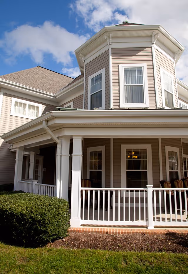 Front exterior of a light-colored two-story building with a covered wraparound porch and white railing under a blue sky.