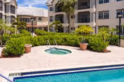 Outdoor view of a senior living facility courtyard featuring a swimming pool and a hot tub surrounded by potted plants and greenery, with a multi-story building in the background under a partly cloudy sky.