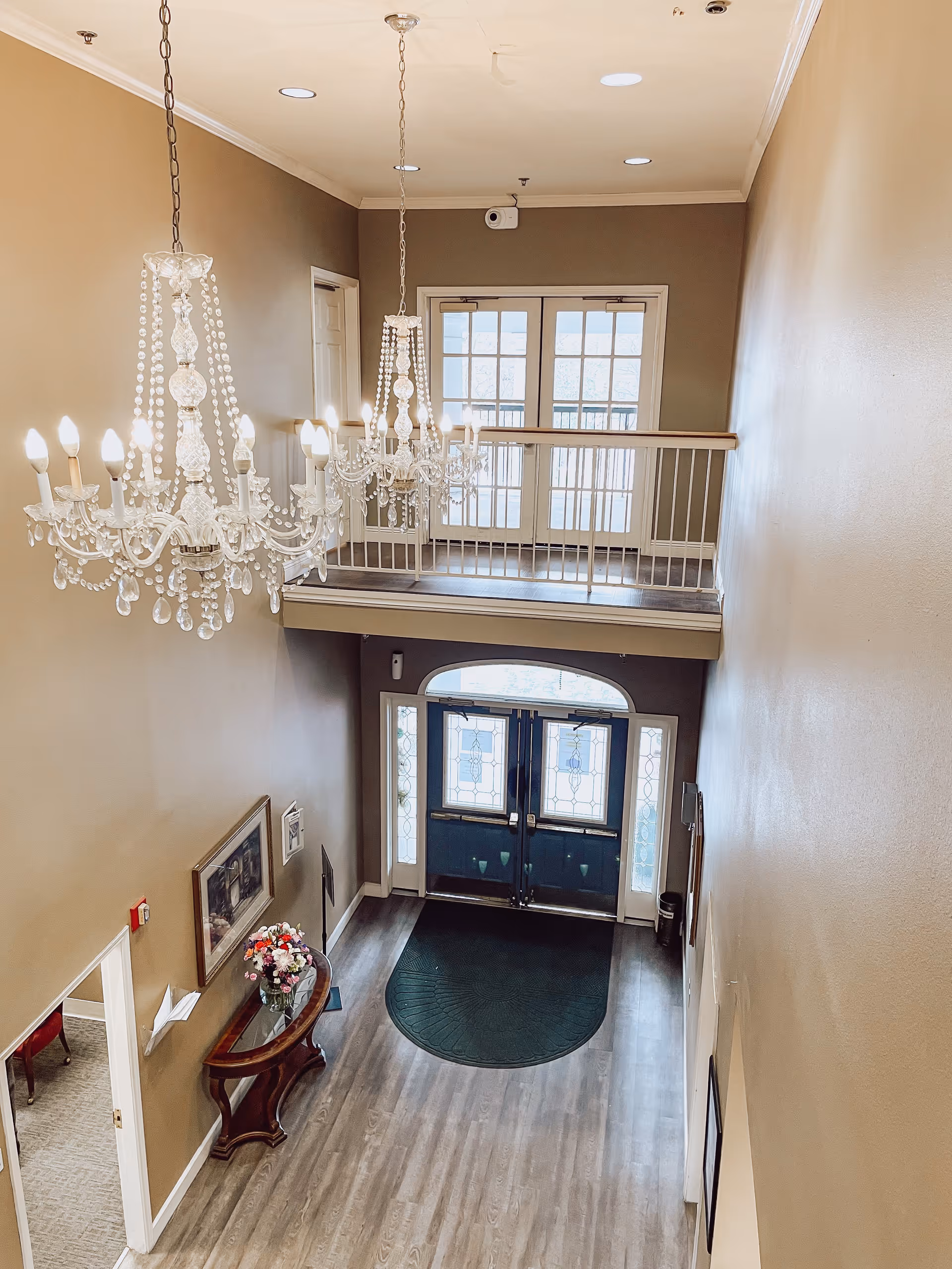 View from above of an elegant entryway with double glass doors featuring decorative glass panels. Two large crystal chandeliers hang from the ceiling. There is a small wooden table with a flower arrangement and a framed picture on the wall. The floor is wood with a dark green mat near the doors, and there is a balcony railing on the upper level overlooking the entryway.