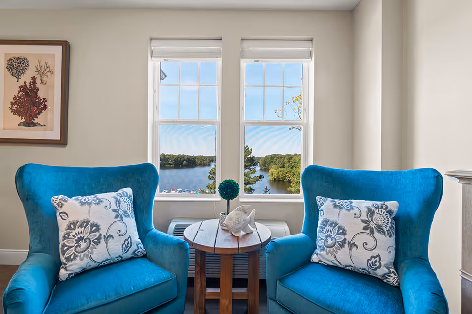 Two teal armchairs with patterned pillows flanking a small round table in front of a window overlooking a lake.