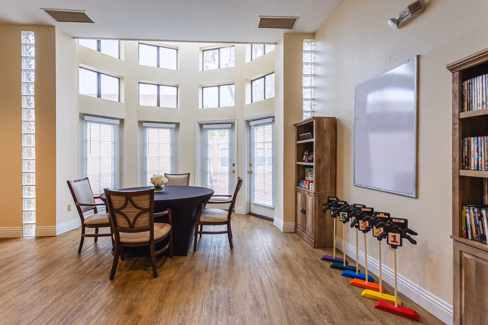A bright room with large windows and glass block accents, featuring a round table covered with a black tablecloth surrounded by four wooden chairs with cushioned seats. There are wooden bookshelves on the right side filled with books and games, a whiteboard mounted on the wall, and a set of numbered horse-shaped game pieces lined up on the floor.