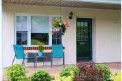 Outdoor patio area with a small table and two teal chairs. A hanging flower basket is suspended above the table, and a potted plant sits on the table. The patio is adjacent to a beige building with a green door and two windows. There are various green shrubs and plants in front of the patio.