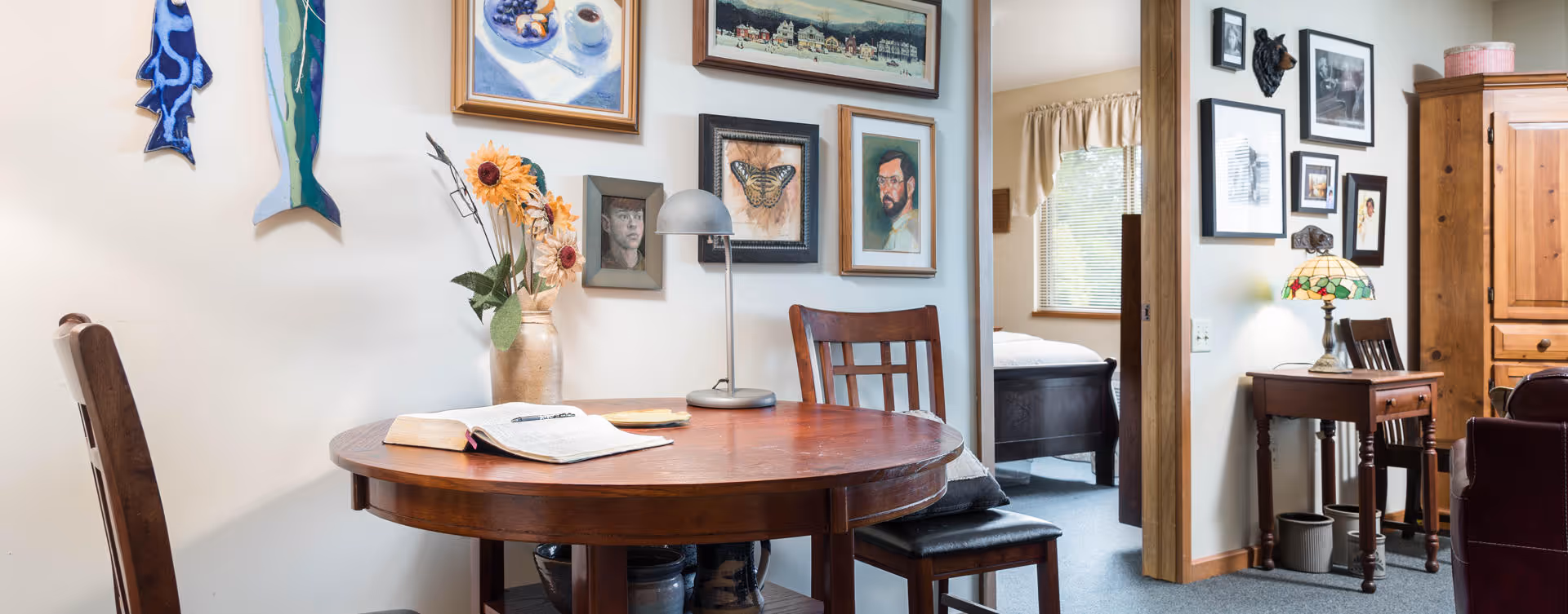 A cozy interior space featuring a round wooden table with an open book and a vase of sunflowers. The walls are decorated with various framed pictures and artwork. To the right, there is a small wooden desk with a Tiffany-style lamp and a wooden armoire. An open doorway reveals a bedroom with a bed and window with curtains.