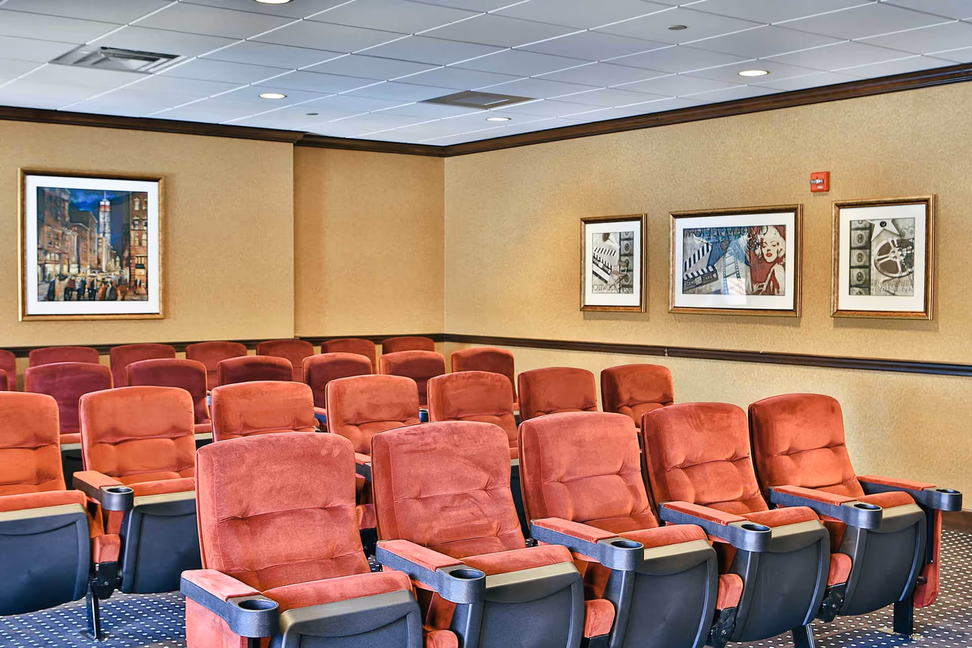 A small theater room with multiple rows of red cushioned seats with black armrests and cup holders. The walls are beige with dark brown trim, decorated with framed artwork including cityscape and film-themed pictures. The ceiling is white with recessed lighting.
