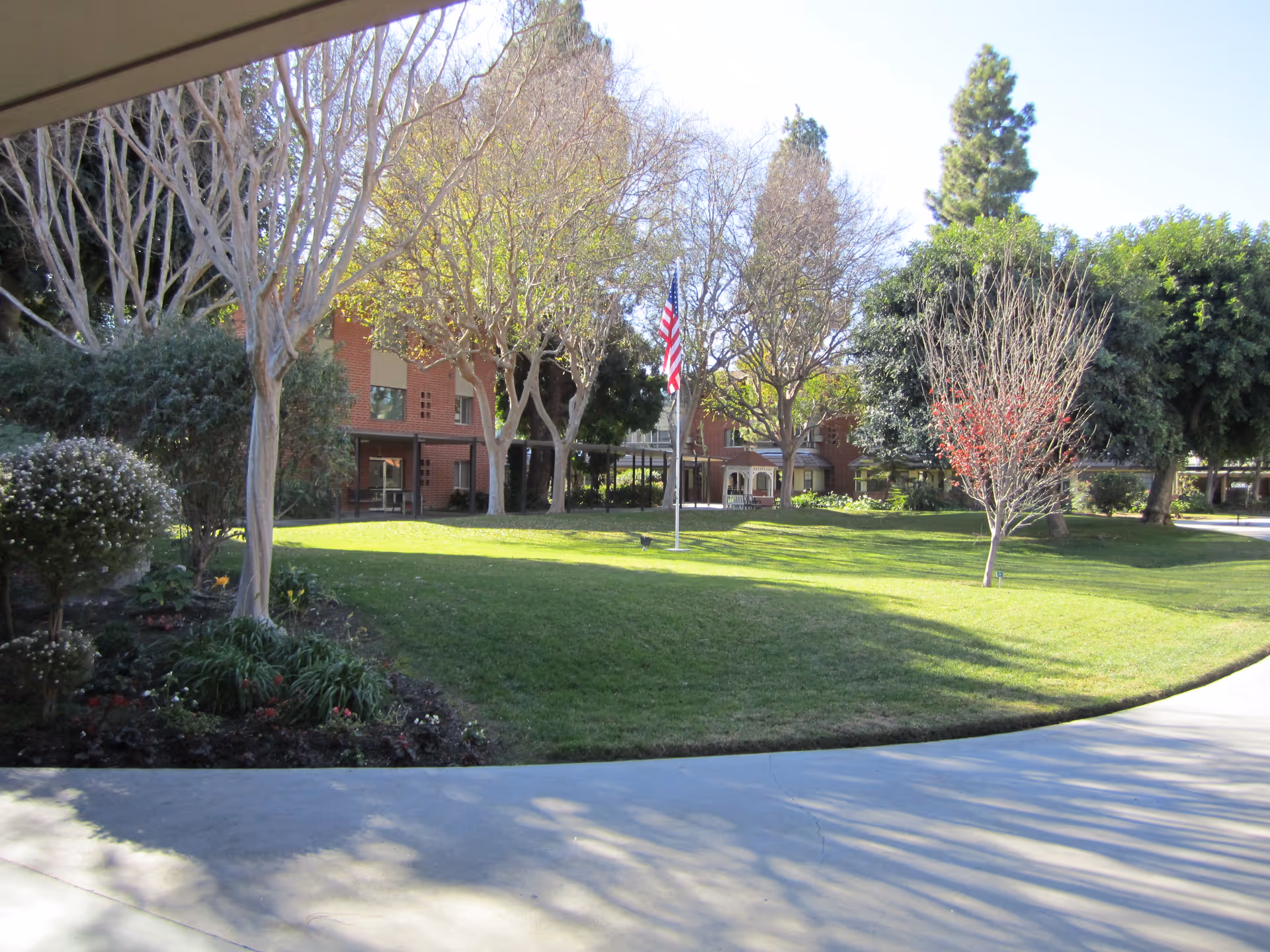 A well-maintained outdoor garden area with green grass, various trees, and shrubs. In the background, there is a brick building with multiple windows and a covered walkway. An American flag is prominently displayed on a flagpole in the center of the grassy area.