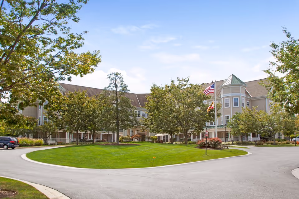 Exterior view of Sunrise of Columbia senior living facility showing a large, multi-story building with beige siding and a green roof. The building is surrounded by well-maintained green lawns, trees, and a circular driveway. Two flagpoles with the American flag and a state flag are visible near the entrance.