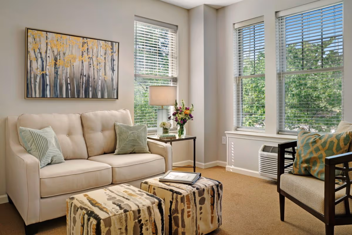 Bright living room with a beige loveseat, two patterned ottomans, an armchair, side table with a lamp and flowers, and large windows with blinds showing greenery outside.