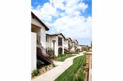 Exterior view of a row of residential buildings with white walls and brown tiled roofs along a paved walkway under a partly cloudy blue sky.