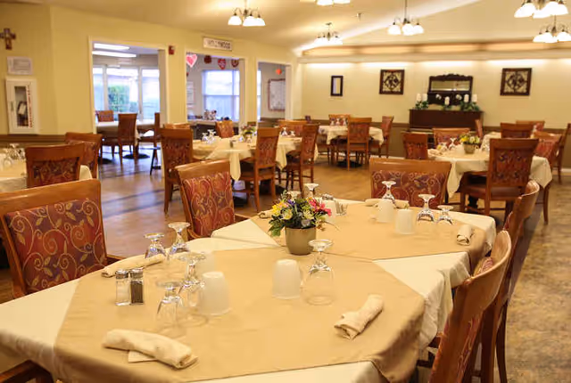 A dining room in a senior living facility with multiple tables covered in beige tablecloths and set with glasses, napkins, and a small flower centerpiece. The room has wooden chairs with patterned upholstery and warm lighting from ceiling fixtures. There are windows and doors visible in the background.