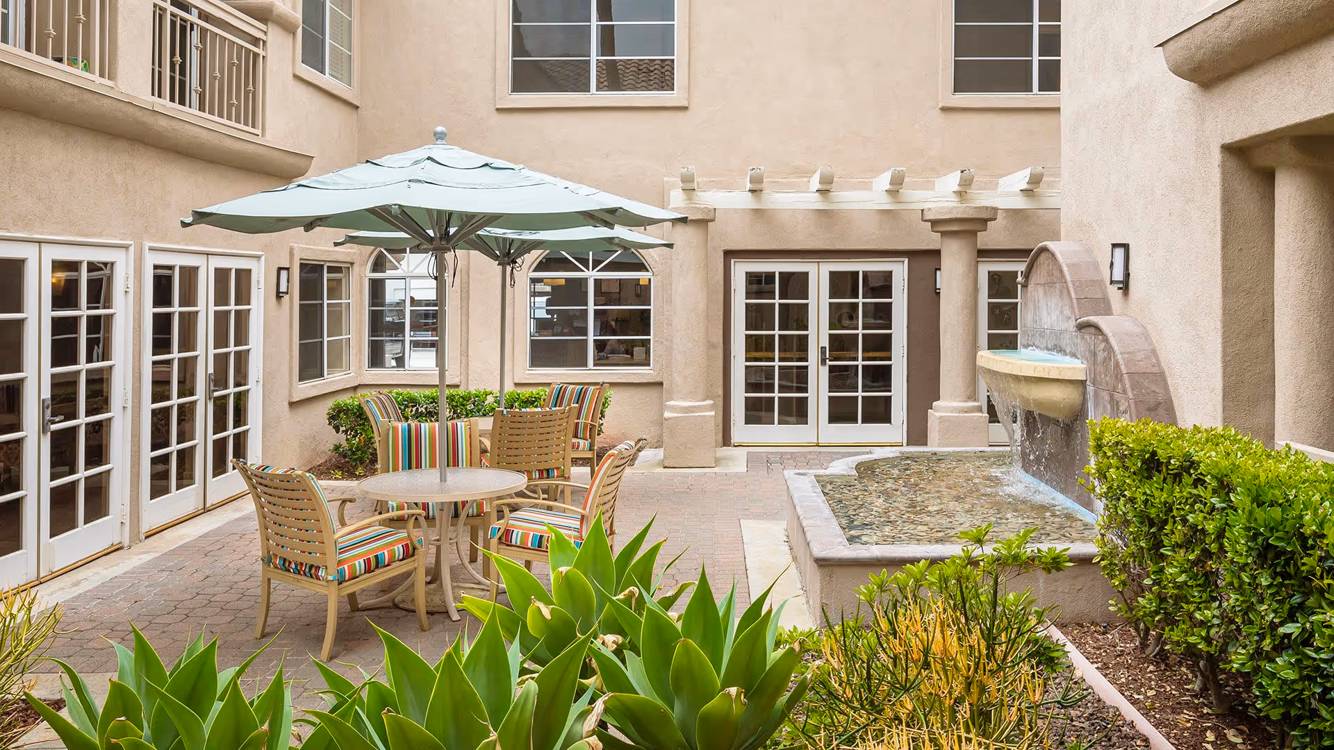 Outdoor courtyard area with a round table and four striped cushioned chairs under a large umbrella. There is a water fountain feature on the right side and green plants surrounding the paved patio. The building walls are beige with multiple windows and glass doors.