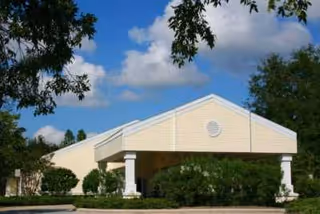 Exterior view of a single-story building with a light yellow facade and white trim, featuring a covered entrance supported by white columns. The building is surrounded by green shrubs and trees under a blue sky with scattered clouds.