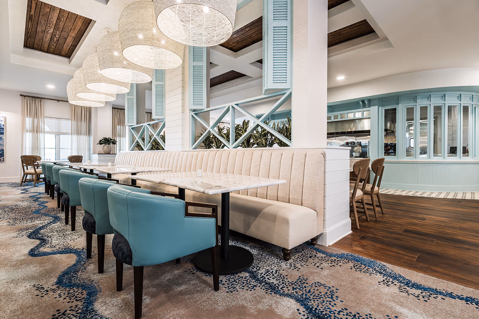 A stylish dining area in a senior living facility featuring a long beige upholstered bench along a wall with multiple marble-top tables and teal cushioned chairs. The space has large hanging woven pendant lights, light blue decorative wooden panels, and a patterned carpet with blue and beige tones. The background shows additional seating and a kitchen area with light blue cabinetry and wooden flooring.