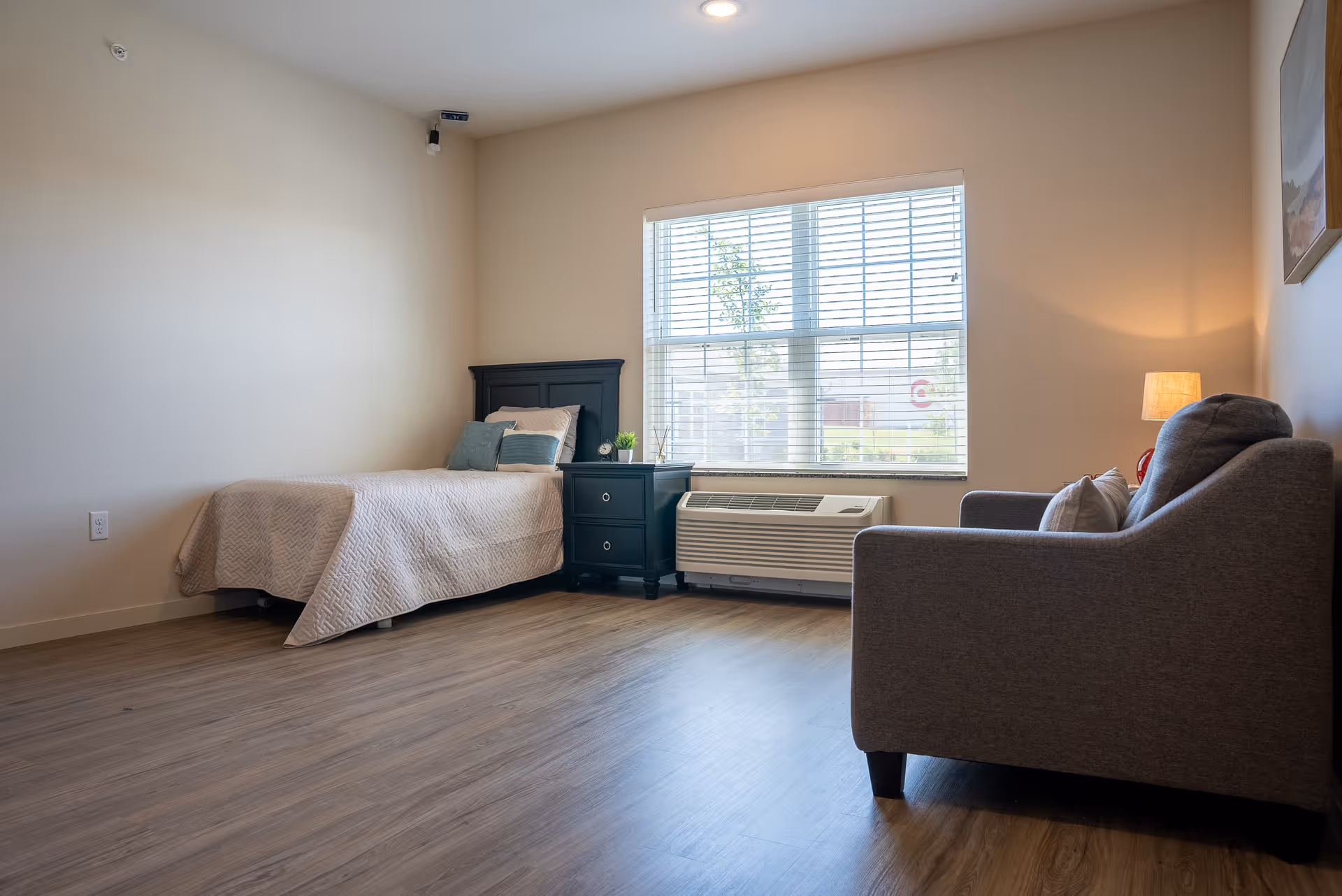 A simple, clean senior living bedroom with a single bed covered in a beige quilt, a black nightstand with decorative items, a large window with blinds, and a gray armchair with a lamp beside it.