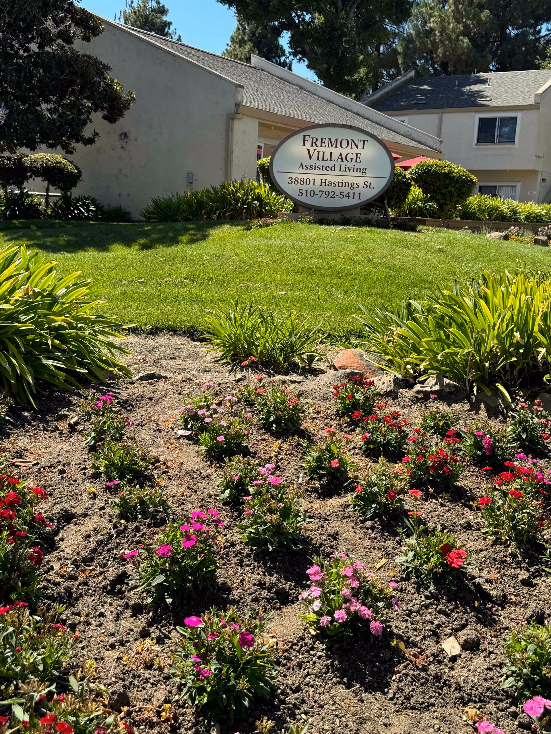 Outdoor view of Fremont Village Assisted Living facility showing a garden with blooming pink and red flowers in the foreground, green grass, bushes, and trees. A sign displays the facility name, address, and phone number in front of a beige building with a sloped roof.