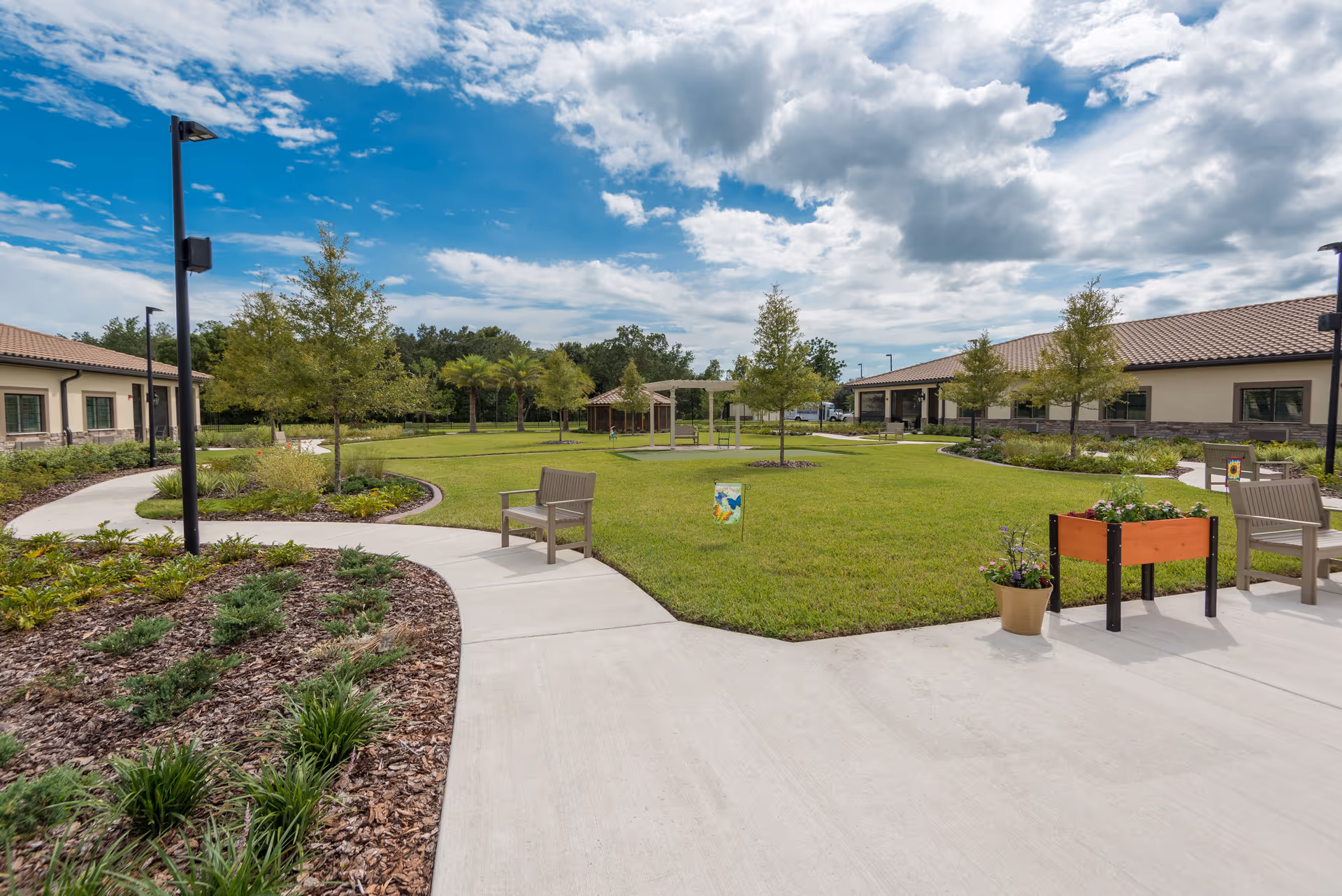 Outdoor courtyard area at Inspired Living Ocoee featuring a well-maintained lawn, several small trees, benches, flower pots, and a paved walkway under a partly cloudy blue sky.