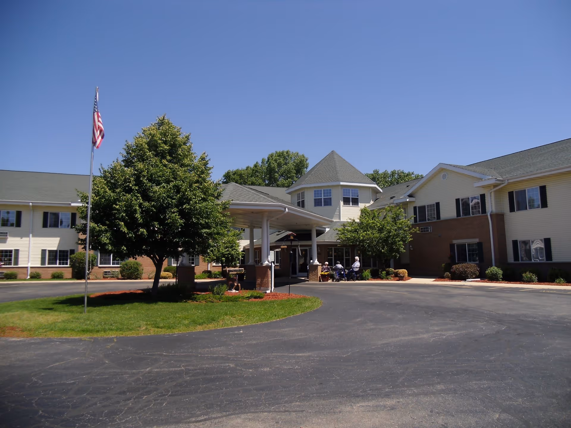 Exterior view of Aster Assisted Living Of Monroe facility on a clear sunny day, showing a large two-story building with beige siding and green roof. There is a covered entrance with a few people sitting near it, a circular driveway, a flagpole with an American flag, and several trees and shrubs in the landscaped area.