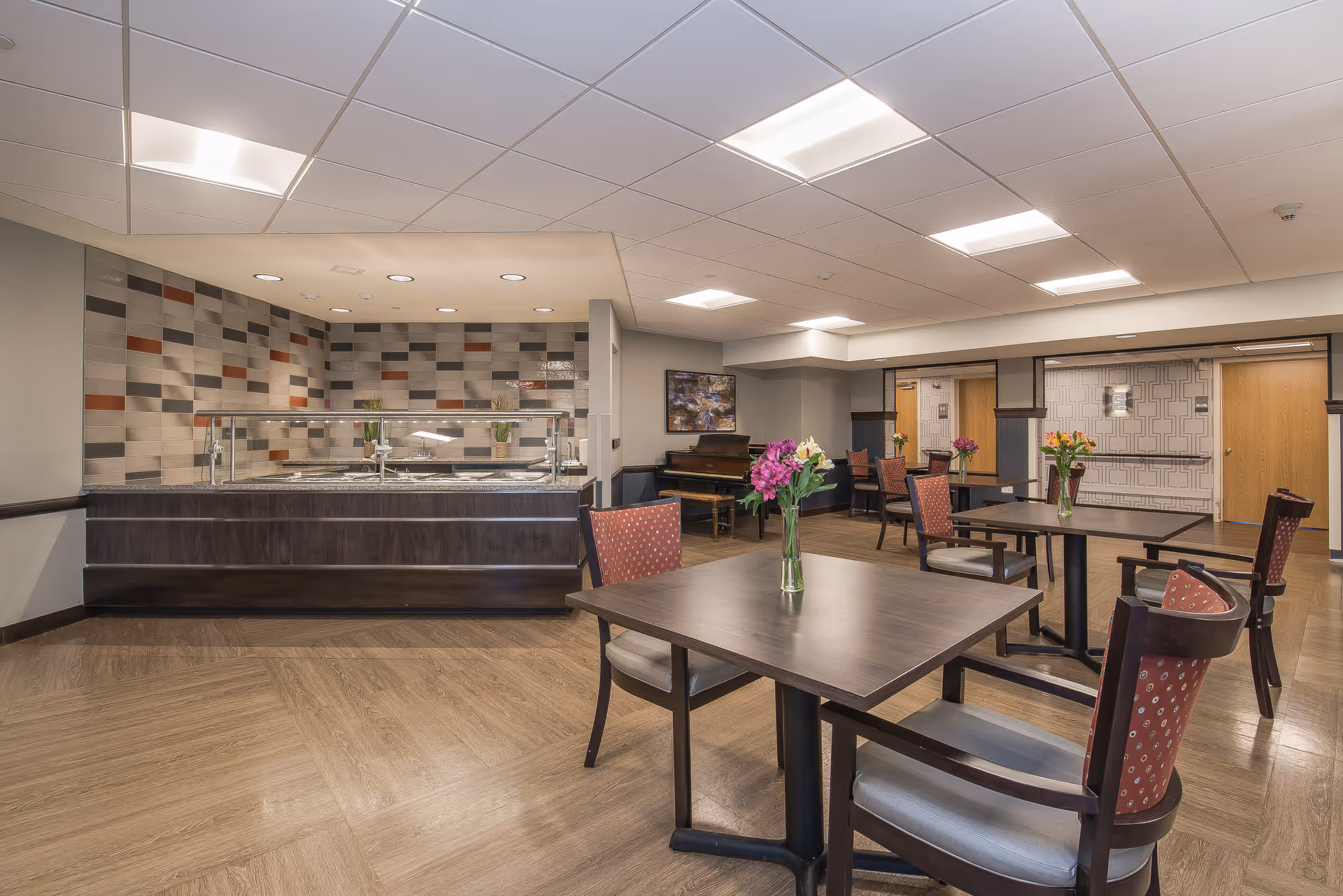 A dining area in a senior living facility featuring several dark wood tables with chairs that have red patterned cushions. Each table has a vase with colorful flowers. In the background, there is a serving counter with a glass sneeze guard and a tiled wall with a geometric pattern. The room has a light-colored ceiling with recessed lighting and wood-patterned flooring.