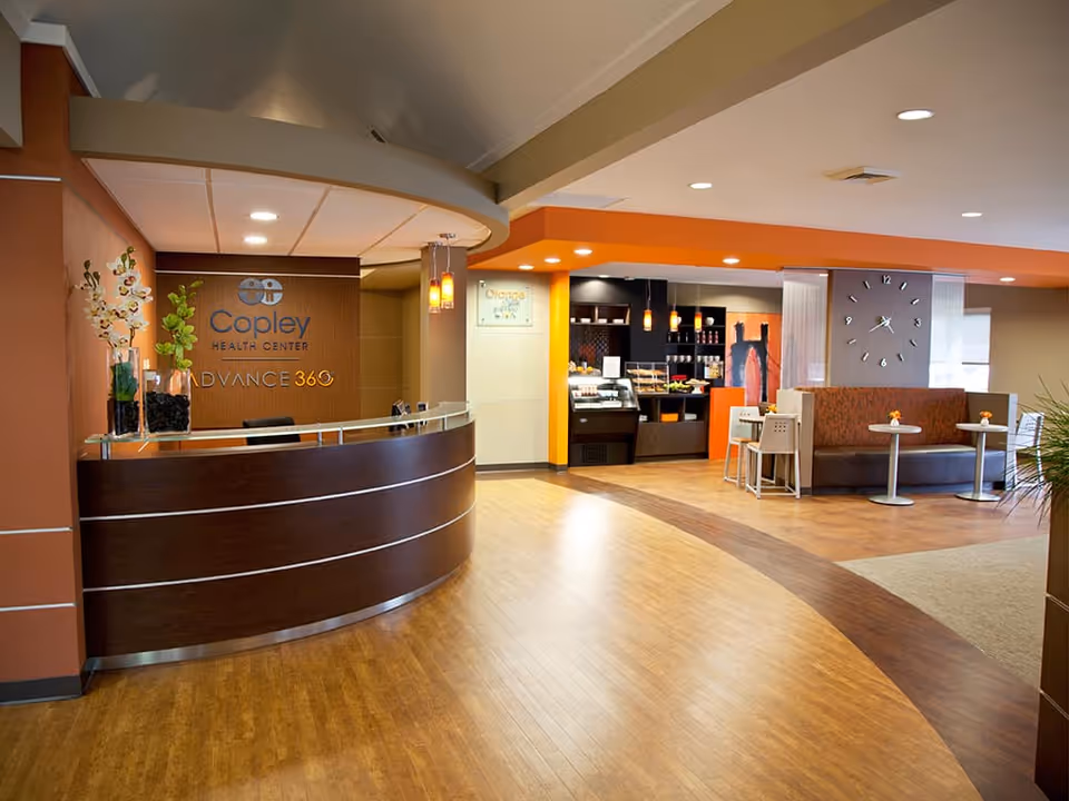 Interior view of a healthcare center reception area with a curved wooden reception desk on the left, decorated with vases of flowers. Behind the desk is a wall with the signage 'Copley Health Center ADVANCE 360'. To the right, there is a small café area with tables, chairs, and a display counter with food items. The floor is wooden, and the ceiling has recessed lighting.